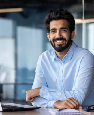 Portrait of happy and successful businessman, indian man smiling and looking at camera, satisfied with achievement results man working inside office building using laptop at work.